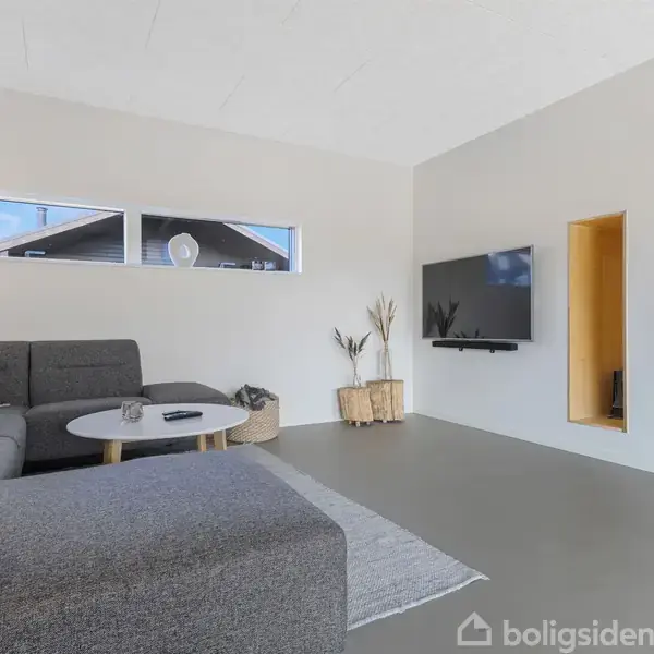 Grå sofa står i stuen ved et rundt sofabord på en lys tæppe. Fjernsynet er vægmonteret, og små vinduer giver lysindfald. Væggen har en indbygget reolniche.