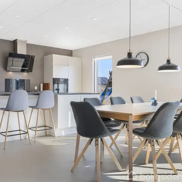 Køkken med moderne spisebord omkranset af otte grå stole. I baggrunden ses et køkkenområde med barstole, køkkenudstyr og store vinduer, der giver udsigt til en terrasse.