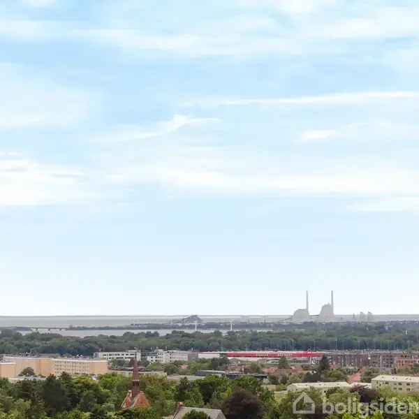 Et kraftværk står i det fjerne, omgivet af en by og grønne træer. En klar, blå himmel breder sig over landskabet.