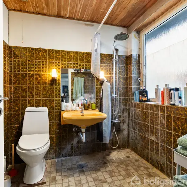 Toilet i en brunflisebeklædt badeværelse, placeret ved siden af en gul vask med spejl. Foran et vindue står diverse plejeprodukter. Rummet har en bruseniche og hvide døre.