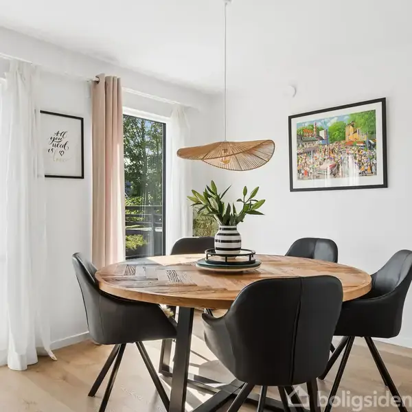 Et rundt træbord med sorte stole står i en lys spisestue. En lampe hænger over bordet, og der er planter samt vægkunst i rummet. Udenfor er der træer.