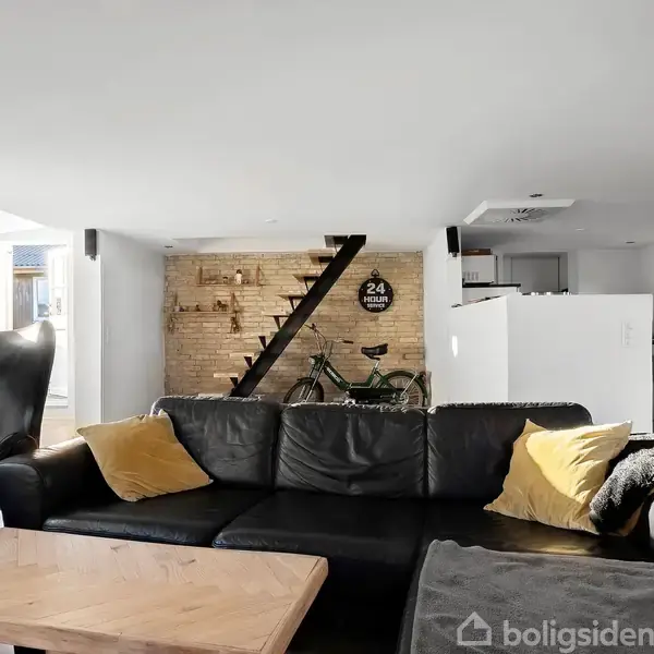 Sort lædersofa med gule puder står foran et træbord i en moderne stue. En cykel er placeret ved en murstensvæg, og en trappe leder til en anden etage. Klokke viser "24 Hours".