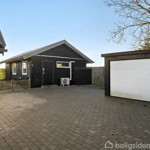 En hvid bolig med sort tag står i en gårdhave. Terrassen har havemøbler og grill. Omkring huset er en velholdt, grøn græsplæne.