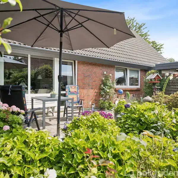 En terrasse med et spisebord og parasol står foran et rødt murstenshus. Farverige blomster omgiver området i en frodig have.