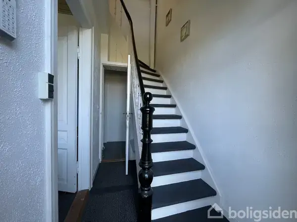 En trappe med hvidmalede trin og sort gelænder fører opad i en lys gang. Døre åbner sig til rum på venstre hånd, med vægdekorationer langs trappevæggen.