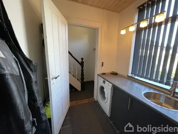 Vaskemaskine står stille i et bryggers ved siden af en vask på en bordplade. Til venstre hænger jakker, mens en åben dør fører til en trappeopgang.