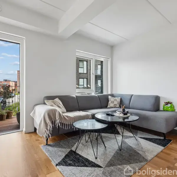 En moderne stue med en grå hjørnesofa, placeret på et tæppe. Rummet har parketgulve, med et glasbord foran sofaen. En åben altandør viser udsigt til byen udenfor.