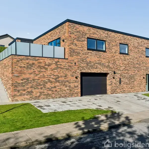 Moderne murstenshus med fladt tag, garage, og stor terrasse. Huset ligger på en grøn græsplæne i et villakvarter med en vej foran.