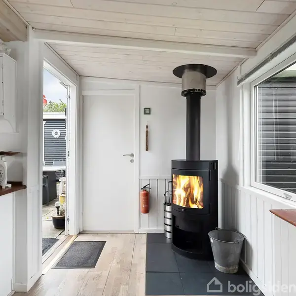Brændeovn i gang emmer af hygge, mens ilden brænder. Rummet har hvide vægge og gulv-til-loft vindue, et åbent indgangsparti og en køkkenbordplade til venstre overfor døren.