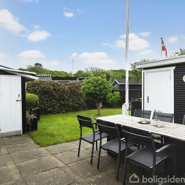 Et havebord med stole står på en terrasse ved siden af et sort skur. Skuret omkranser en græsplæne med buske og et flag i baggrunden.
