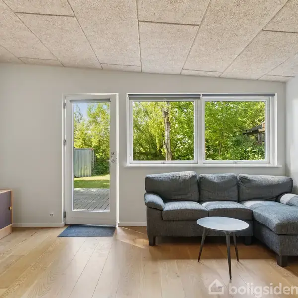 Sofa står i en lys stue ved siden af et lille rundt bord, foran store vinduer og en terrassedør, som åbner ud til en have med grønne træer.
