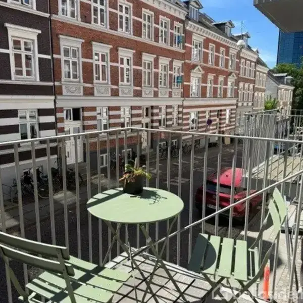 En lille altan med et grønt bord og en plante står ud til en byvej med brostenslagte bygninger og parkerede cykler. En rød bil ses på vejen.