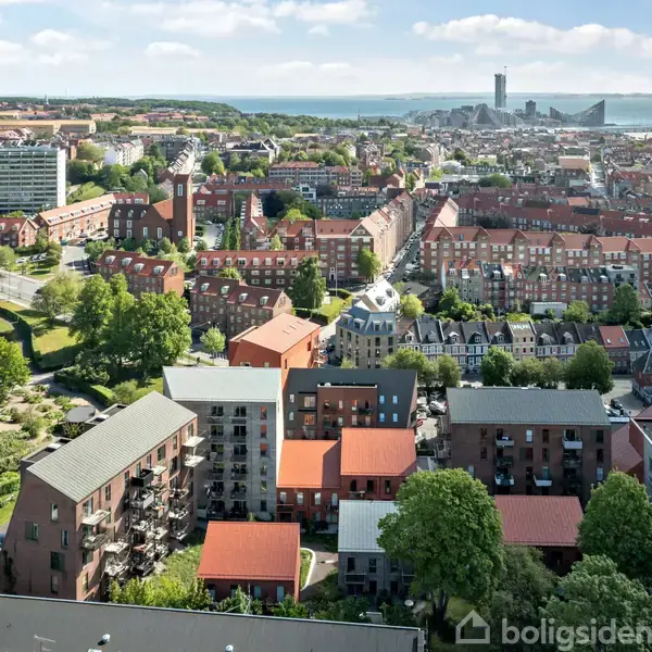 Boligblokke ligger tæt på hinanden med røde tage; omgivende bylandskab og grønne træer. I baggrunden ses en havnefront med moderne bygninger og havet i horisonten.