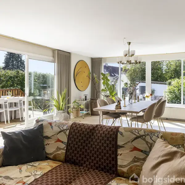 En sofa med mønstret betræk står i en lys stue, fyldt med planter og dekorationer, ved siden af en spisebord. Store vinduer og en udendørs terrasse ses i baggrunden.