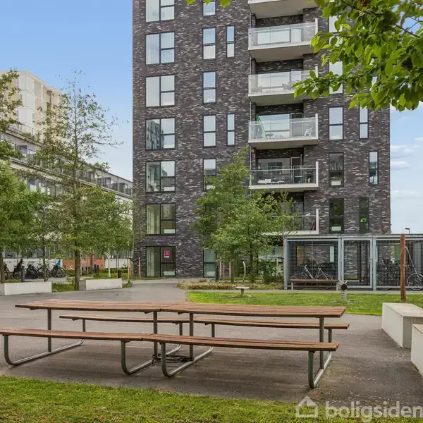 En lang bænk står på en asfaltplads foran et moderne lejlighedskompleks med altaner. Omkring bænken er der græs og træer, hvilket skaber en grøn bymiljø.