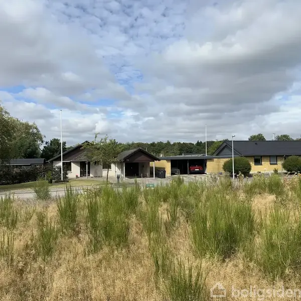 To huse står side om side med velholdte forhaver. Baggrund er markeret af skov og himmel med skyer. Foran huse er en mark med højt græs og ukrudt.