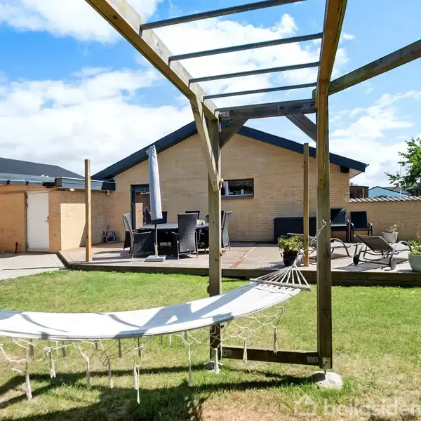 Hængekøje hænger fra en træpergola i en have. I baggrunden ses en terrasse med havemøbler og en gul murstensbygning under en blå himmel.