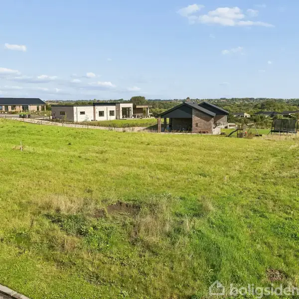 En græsmark strækker sig ud i forgrunden, mens to moderne huse står stille i det fjerne. Omgivet af en grøn, landlig baggrund under en blå himmel med spredte skyer.