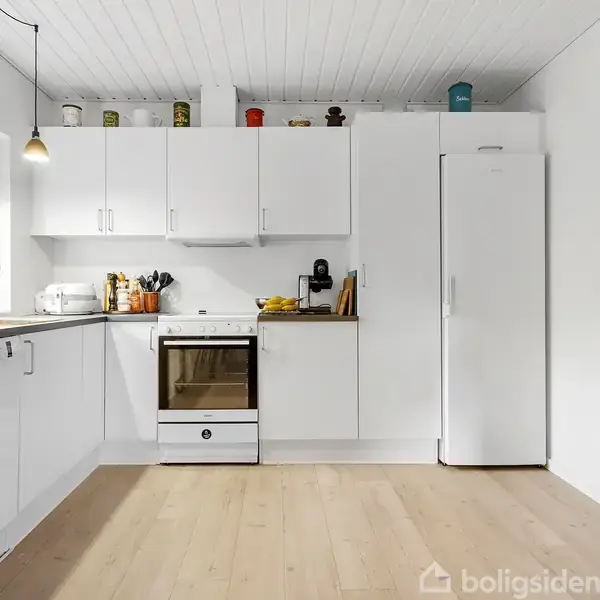 Et hvidt køkken med skabe og en ovn i midten. En opvaskemaskine står til venstre. Vinduer med gardiner lader naturligt lys strømme ind. Trægulvet skaber en varm atmosfære.