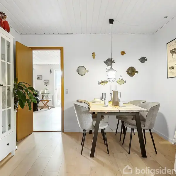 Et spisebord med seks stole er opstillet i en spisestue. Væggen er dekoreret med fiskedekorationer, og der hænger en lampe over bordet. En hvid bogreol står i hjørnet.