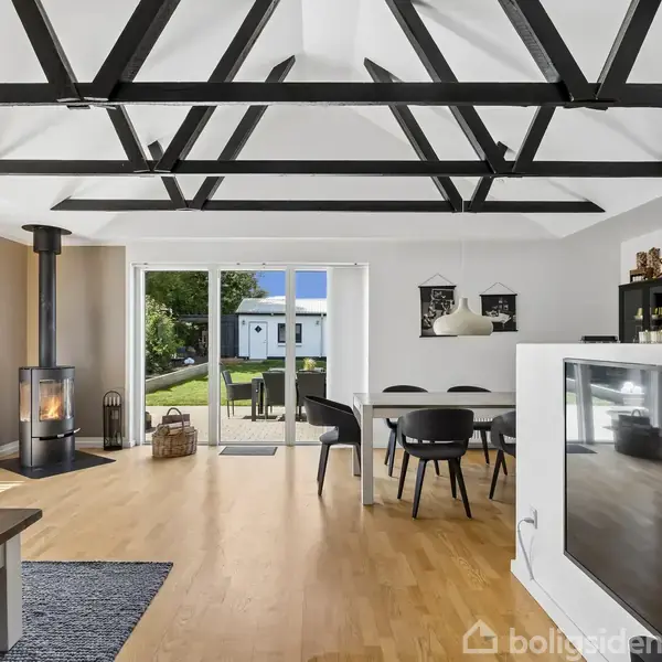 Stue med træbjælker i loftet åbner til have gennem skydedør. Brændeovn står i hjørne, og et moderne spisebord med sorte stole dominerer rummet.