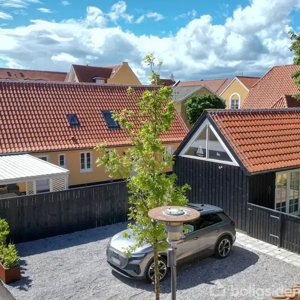 Bil holder parkeret på grusvej foran sort hus med rødt tag. Omgivet af træer og andre huse med orange tage under en blå himmel med skyer.