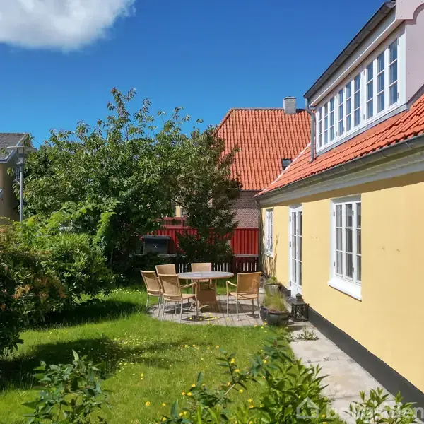 Gult hus med rødt tag står i en solrig have. Rundt havebord med stole er omgivet af grønbeplantning. Klart blå himmel og en anden bygning i baggrunden.