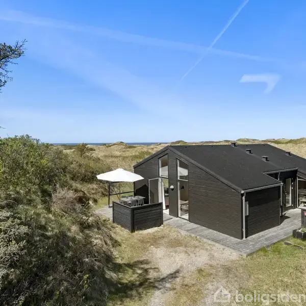 Et moderne sommerhus står midt i klitlandskabet med en terrasse og parasol ved siden af. Omkring huset er der sandklitter og buske under en klar blå himmel.
