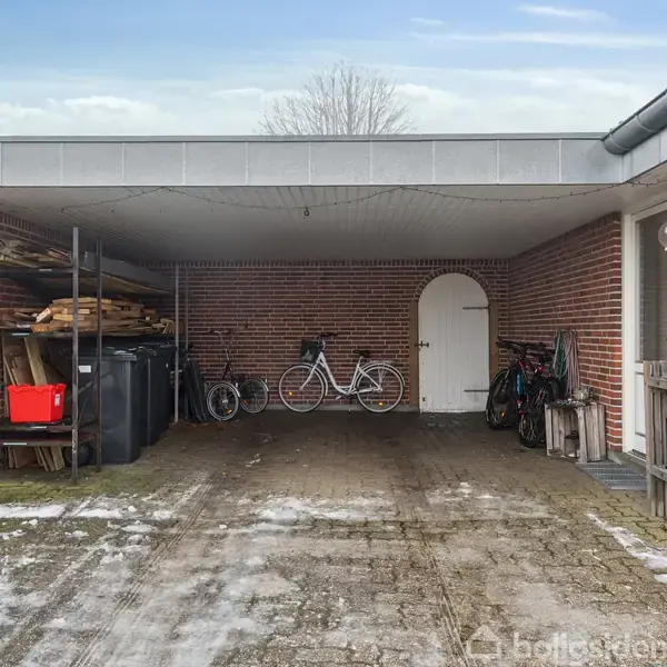 Carport med to cykler står på en flisebelagt indkørsel. Ved siden af er der hylder med brænde, skraldespande og en lukket dør. Snedækket jord omgiver området.