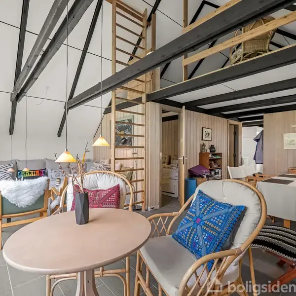 Et rummeligt stueområde med loft til kip og synlige sorte bjælker. Der er en lille rund bord med stole og farverige puder. I baggrunden ses en trappe til en hems.