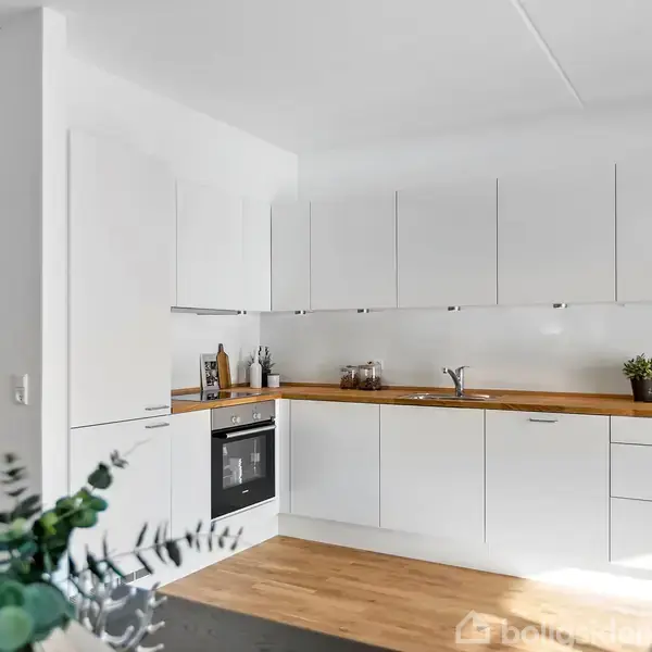 Et moderne køkken med hvide skabe og en træbordplade. En ovn er indbygget i skabet til venstre. Planter og køkkenredskaber står på bordpladen. Sollys lyser rummet op.