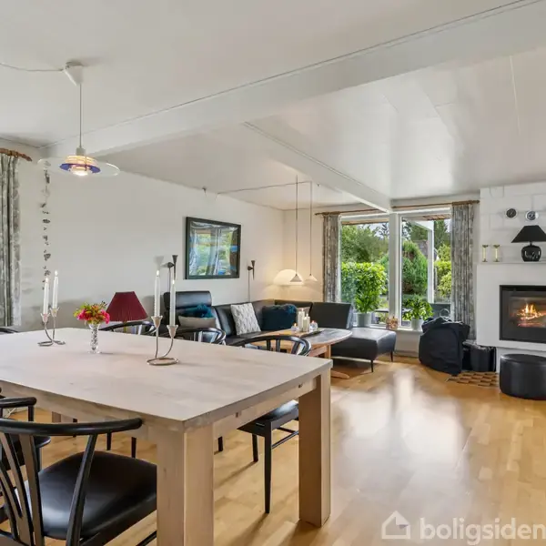 Et lyst rum med spisebord i træ og sorte stole, placeret i nærheden af en sofa med pejs i baggrunden. Store vinduer slipper lys og udsigt til haven ind.