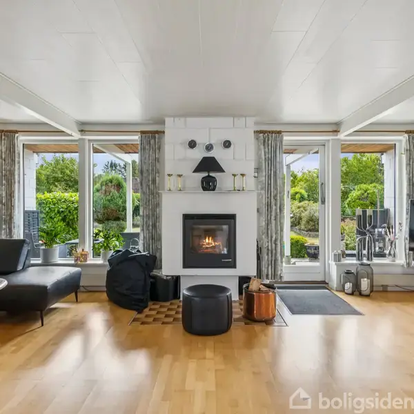 Pejs i funktion, omgivet af moderne stueindretning med lædermøbler, træbord og stort fladskærms-TV. Gennem store vinduer ses en velplejet have, som giver et lyst og indbydende miljø.