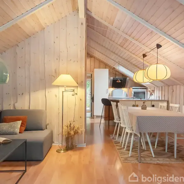 Gråt sofaarrangement i træbeklædt stueetage med skrå vægge. Lampe belyser rummet. Til højre står et spisebord med stole og hængende lamper over.