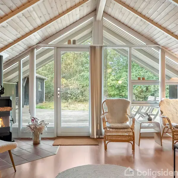 Pejsen er i brug i en hyggelig stue med skrå loft og store vinduer. To kurvestole står ved siden af, og udenfor ses en terrasse og grønne træer.