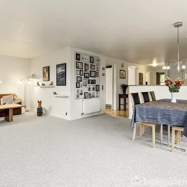 En lys stue indeholder et spisebord med fire stole og en vase med blomster. I baggrunden står en sofa med lamper og hylder. Væggene er dekoreret med billeder.
