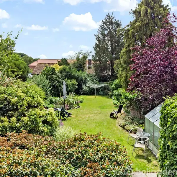 En grøn have med græsplæne og forskellige buske og træer. Der er en parasol og havestole. Omkring haven er der huse og en klar himmel med skyer i baggrunden.