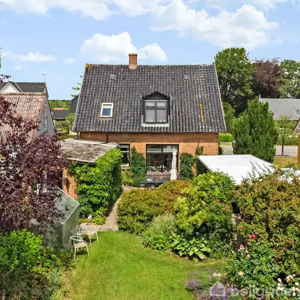 Et rødt murstenshus med mørkt tag står i en frodig have med buske og blomster. Til venstre ses et drivhus og havemøbler. Nabohuse omgiver det fredelige område.