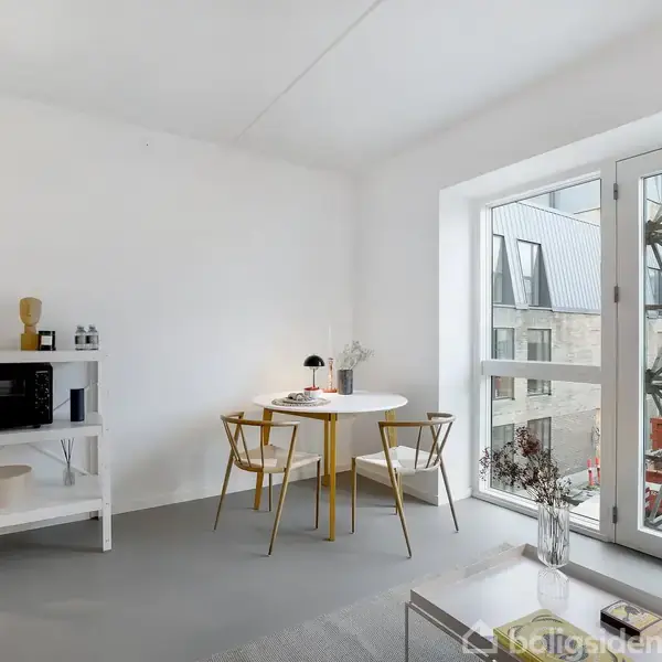 Et rundt bord med to stole står ved et stort vindue, der giver udsigt over en byggeplads. Til venstre ses en hvid reol med en mikroovn og dekorationer.