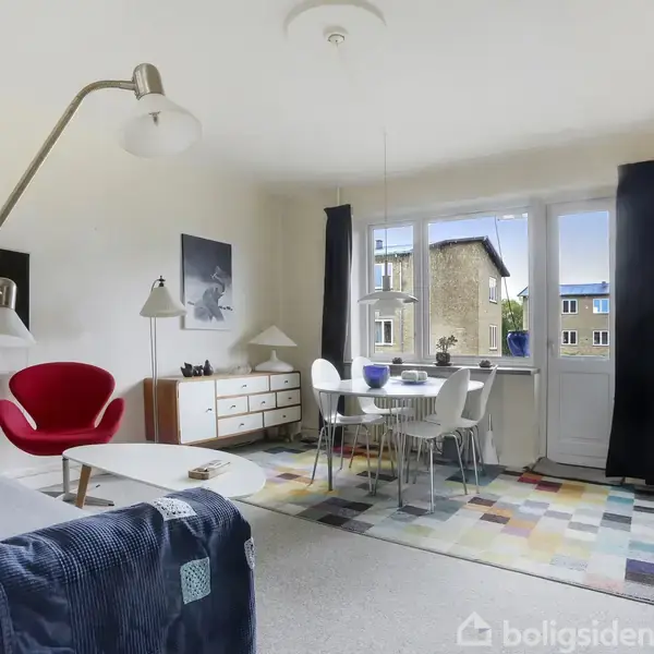 En lys stue med moderne møbler, herunder en rød stol, sofa, og spisebord. Store vinduer giver udsigt til en bygning udenfor. Rummet har en farverig tæppe på gulvet.