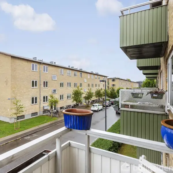 Blomsterkrukker står på altanen; bygningen er af lys mursten med grønne elementer. Vejen nedenfor er omkranset af græs og træer. Det er en boligområde med køretøjer parkeret langs vejen.