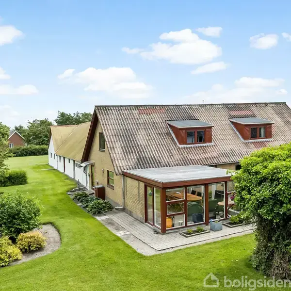 En stor, rødtagset villa med flere kviste står stille i en velholdt have med buske og træer, omkranset af frodige græsarealer, under en klar blå himmel med spredte skyer.