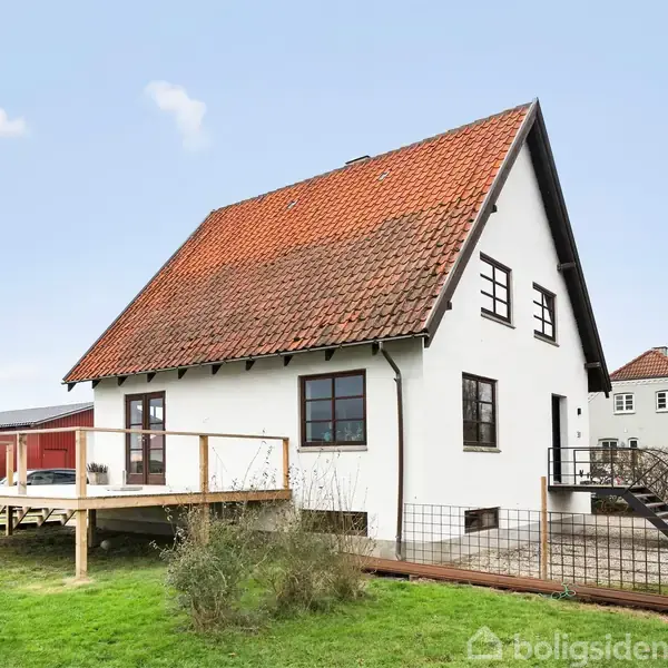 Et hvidt hus med rødt tegltag står på en græsplæne. Huset har en træterrasse i forgrunden, mens skyfri himmel og nabohuse er synlige i baggrunden.