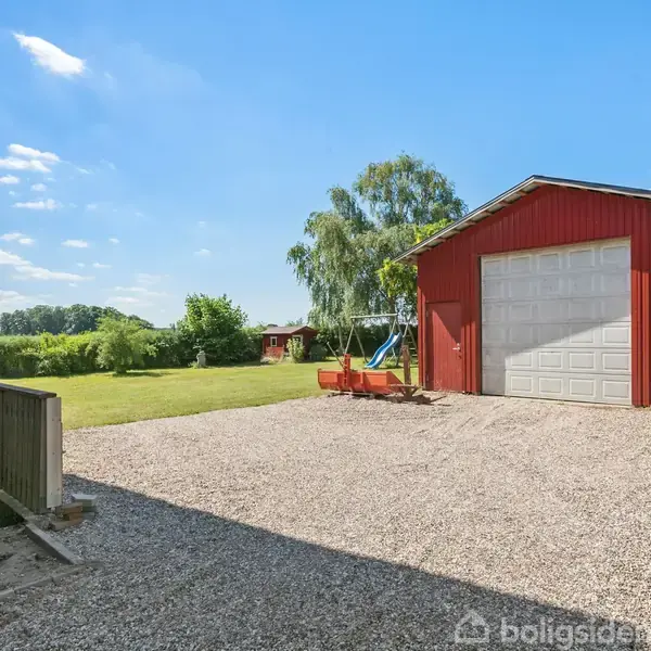 Rødt skur med hvid port står på grusplads. Ved siden af er en gynge, og baggrunden viser en grøn have og træer under en blå himmel.