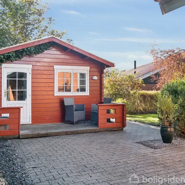 En rød træhytte står stille på en flisebelagt gårdhave omkranset af buske. Hytten har en hvid dør og vindue med skodder. En stol og et bord står foran hytten.