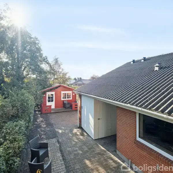 Garagen er tilknyttet et rødt murstenshus med sort tag, blandt træer og buske på en solrig dag. En mindre rød bygning ses i baggrunden ved enden af en stenbelagt indkørsel.