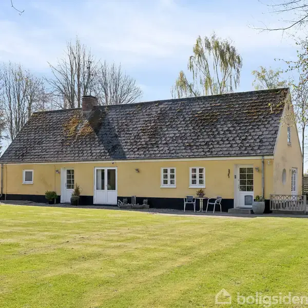 Gult hus med skråt tag står stille på en velplejet græsplæne; omgivet af træer og buske under en blå himmel.