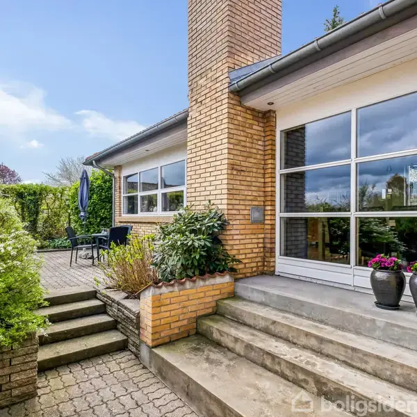 Hus med gul murstensfacade, stor glasdør og blomsterkrukker på trappen. En terrasse med bord og blå paraply ved siden af, omkranset af grønne buske.