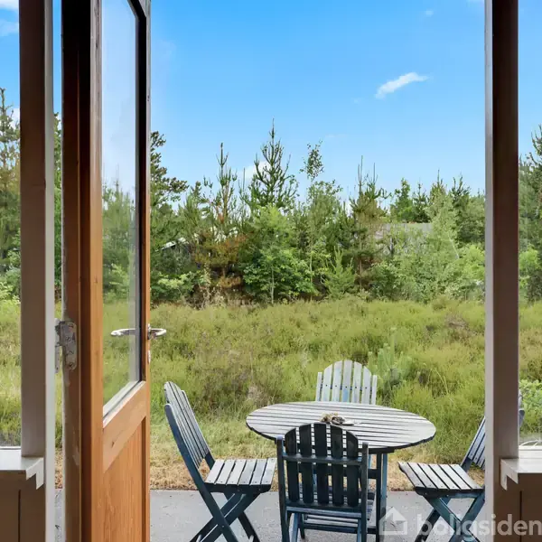Dør står åben mod en terrasse med et rundt bord og fire stole. I baggrunden ses en frodig skov med grønne træer under en blå himmel.