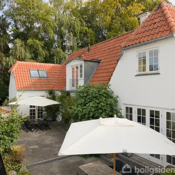 Huset har hvid facade og rødt tegltag, omgivet af grønne træer. Terrassen har møbler og parasoller. Vinduer med sprosser lader lys ind i rummet.